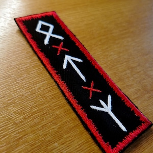 May include: Black fabric patch with a red border. The patch features four white embroidered symbols, including a cross, an arrow, and two runes.