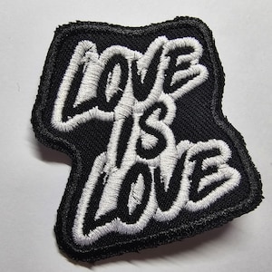 May include: A black embroidered patch featuring the phrase "LOVE IS LOVE" in white, cursive-style lettering. The patch has a black border and is designed to be sewn or ironed onto clothing or fabric.