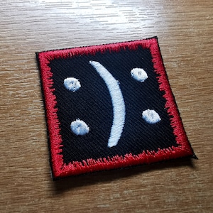 Può includere: Un cerotto in tessuto nero con un viso triste ricamato in bianco e un bordo cucito rosso.