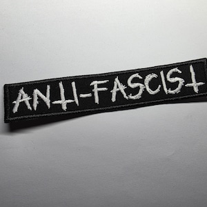 Puede incluir: Parche rectangular negro con la inscripción bordada en blanco "ANTI-FASCIST". El parche tiene un diseño sencillo, con un mensaje claro.
