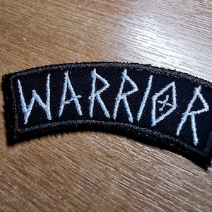 May include: A black fabric patch with white embroidered lettering that reads "WARRIOR".