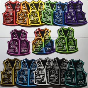 May include: A selection of embroidered vest patches in a range of colours, such as red, orange, yellow, green, blue, purple, and black. Each patch has a black base with white and coloured details, including the letter "X", circles, and geometric shapes. The patches are arranged in rows.