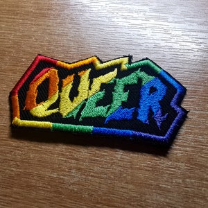 May include: A rainbow-colored embroidered patch with the word "QUEER" in a bold, stylized font.