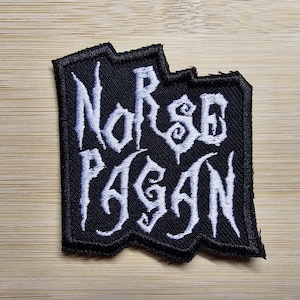 May include: Black embroidered patch with the words "Norse Pagan" in white, stylized lettering. The patch has a jagged edge and is set against a wooden background. The patch is likely for clothing or accessories.