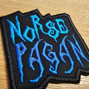 May include: A black embroidered patch with the words "Norse Pagan" in blue lettering. The patch has a jagged edge.
