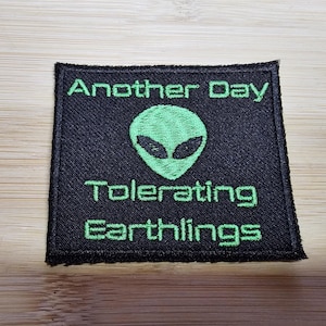 Può includere: Toppa in tessuto nero con testo ricamato in verde che recita "Another Day Tolerating Earthlings". Un viso alieno verde è ricamato al centro della toppa.