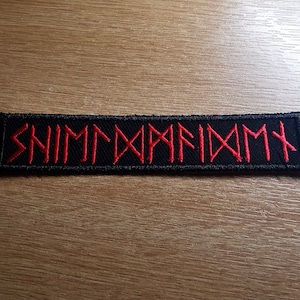 May include: A rectangular black patch with red embroidered runes. The runes spell out a word or phrase. The patch is designed to be sewn or ironed onto clothing. The background is a light brown wooden surface.