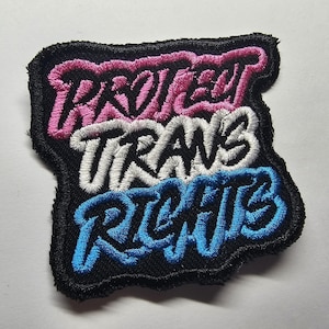 Op de afbeelding: Geborduurde patch met de woorden "PROTECT TRANS RIGHTS" in roze, wit en blauw, op een zwarte achtergrond. De patch heeft een onregelmatige vorm en is bedoeld om op kleding of accessoires te worden genaaid.