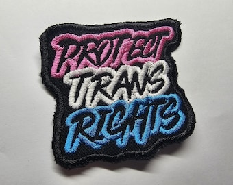 Puede incluir: Parche bordado con las palabras "PROTECT TRANS RIGHTS" en rosa, blanco y azul, sobre un fondo negro. El parche tiene una forma irregular y está diseñado para ser cosido en ropa o accesorios.