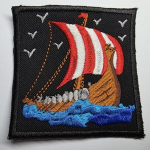 May include: Embroidered patch featuring a Viking ship with a red and white striped sail, sailing on blue waves. The ship is brown with a small red flag. White birds fly in the black sky. The patch is square with a black border.