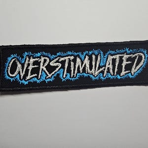 Può includere: Toppa ricamata nera con la scritta bianca "OVERSTIMULATED", bordata di blu. La toppa è rettangolare e ha un bordo nero. Il testo è in un carattere audace e stilizzato.