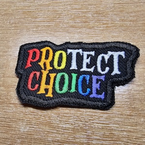 May include: A black embroidered patch with the words "PROTECT CHOICE" in rainbow colors. The words are in a bold, stylized font.