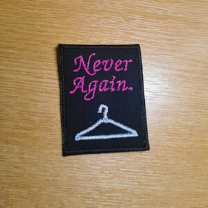 Puede incluir: Parche bordado negro con texto rosa que dice "Never Again." Un colgador blanco está bordado debajo del texto.