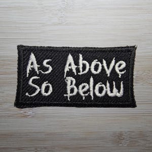 Op de afbeelding: Zwarte geborduurde patch met witte tekst die "As Above So Below" luidt.