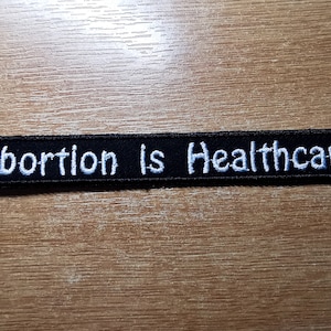 Könnte beinhalten: Schwarzer gestickter Aufnäher mit weißem Text, der "Abortion is Healthcare" lautet.