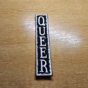 Könnte beinhalten: Schwarzer Stoff-Patch mit weißen gestickten Buchstaben, die vertikal "QUEER" buchstabieren.
