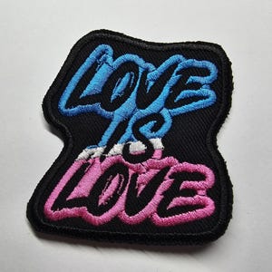 May include: Embroidered patch featuring the phrase "LOVE IS LOVE" in blue and pink script on a black background. The patch has a black border and a white accent between the words.