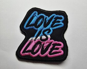 Puede incluir: Parche bordado con la frase "LOVE IS LOVE" en escritura azul y rosa sobre un fondo negro. El parche tiene un borde negro y un acento blanco entre las palabras.