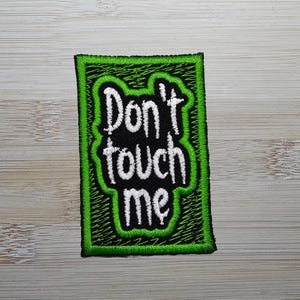 May include: Embroidered patch with the words "Don't touch me" in white, outlined in black, on a black background. The patch has a green border with a zig-zag stitch.