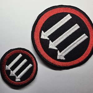 Pode incluir: Dois patches bordados circulares. Cada um tem um fundo preto com três setas brancas apontando para baixo dentro de um anel vermelho. Os patches são de tamanhos diferentes, o maior está à direita.