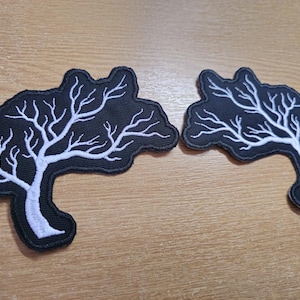 May include: Two black embroidered tree patches with white branches, displayed on a wooden surface. The tree design features a curved trunk and detailed branches. Each patch is approximately 8 cm in width.