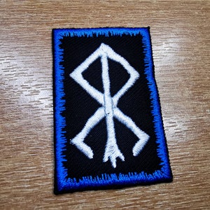 May include: White embroidered symbol on a black patch with a blue border. The symbol is a stylized rune with a central vertical line and four diagonal lines extending from it.