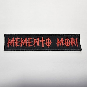 May include: Black rectangular patch with the words "MEMENTO MORI" embroidered in red, gothic-style lettering. The patch has a black border and is likely designed to be sewn onto clothing or accessories.