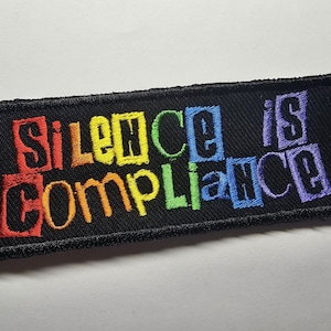 Könnte beinhalten: Schwarzer, rechteckiger, gestickter Aufnäher mit dem Schriftzug "SILENCE IS COMPLIANCE" in Regenbogenfarben. Der Text ist fett und blockartig, und der Aufnäher hat einen schwarzen Rand.