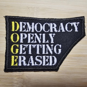 May include: Black patch with yellow letters spelling out "Democracy Openly Getting Erased".