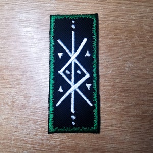 May include: A black and white embroidered patch with a green border. The patch features a white symbol with a vertical line and two diagonal lines intersecting it. The symbol is surrounded by four white triangles and three white dots.