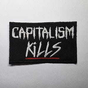Op de afbeelding: Zwart rechthoekig embleem met wit geborduurde tekst "CAPITALISM KILLS" met een rode lijn onder het woord "KILLS". De patch heeft een zwarte rand, geschikt voor kleding of accessoires.