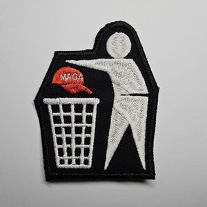 Puede incluir: Parche bordado que representa una figura de palo tirando una gorra de béisbol roja con el texto "MAGA" a una papelera. El parche tiene un fondo negro y costuras blancas.