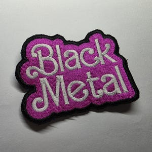 May include: Embroidered patch with the words "Black Metal" in white script on a purple background. The patch has a black border and is likely for clothing or accessories.