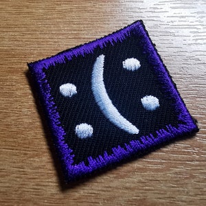 Puede incluir: Un parche de tela negro con una cara triste bordada en blanco. El parche tiene un borde morado.