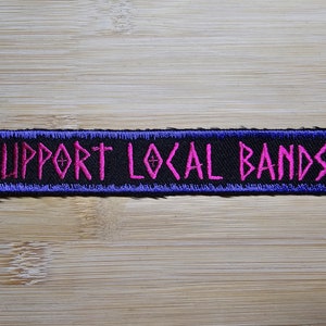 Op de afbeelding: Een zwarte geborduurde patch met een blauwe rand. De tekst "SUPPORT LOCAL BANDS" is roze.