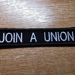May include: A rectangular black patch with white embroidered text that reads "JOIN A UNION". The patch is likely made of fabric and has a border. The text is in a sans-serif font.