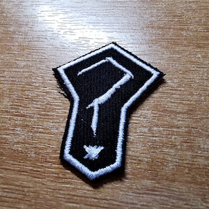 Puede incluir: Un parche bordado en blanco y negro con un signo de interrogación estilizado en forma de pentágono.