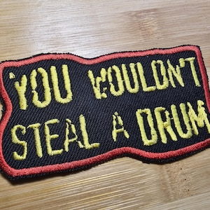 May include: Embroidered patch with the text "YOU WOULDN'T STEAL A DRUM" in yellow thread on a black background. The patch has a red border and is rectangular with a wavy edge.