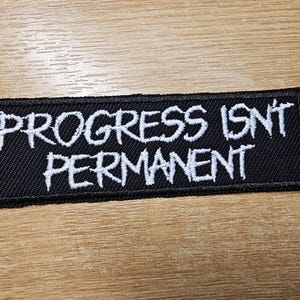 Op de afbeelding: Zwarte patch met witte geborduurde tekst die "Progress Isn't Permanent" luidt.