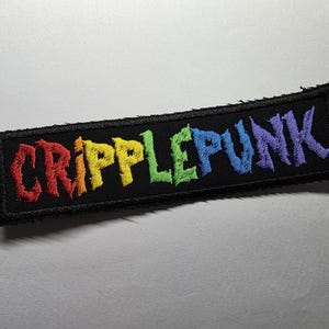 Könnte beinhalten: Ein rechteckiger, schwarzer, gestickter Aufnäher mit dem Wort "CRIPPLEPUNK" in Regenbogenfarben. Der Aufnäher hat einen rauen, unfertigen Rand und ist wahrscheinlich zum Aufnähen auf Kleidung oder Accessoires bestimmt.