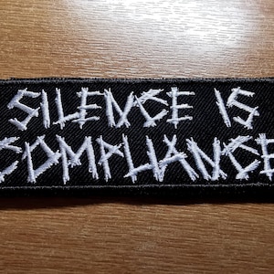 May include: Black embroidered patch with white stitching that reads "Silence is Compliance".