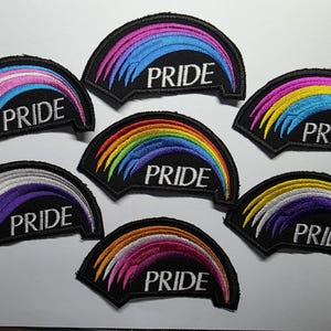 Pode incluir: Sete emblemas bordados semicirculares sobre um fundo preto, cada um apresentando um design de arco colorido e a palavra PRIDE em letras maiúsculas brancas. Os emblemas exibem várias combinações de cores, incluindo arco-íris, rosa, azul e roxo.