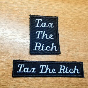 Puede incluir: Dos parches de tela negra con texto bordado en blanco. El parche superior dice "Tax The Rich" en tres líneas. El parche inferior dice "Tax The Rich" en una línea.
