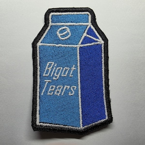 Könnte beinhalten: Gestickter Aufnäher in Form eines Milchkartons mit dem weißen Text "Bigot Tears". Der Karton ist blau mit einer dunkelblauen Seite und einem schwarzen Rand.