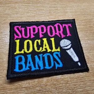 May include: A black embroidered patch with the text "Support Local Bands" in pink, yellow, and blue lettering. A white microphone is embroidered on the right side of the patch.