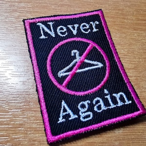 Puede incluir: Parche bordado con las palabras "Never Again" en blanco, con un círculo rosa y un símbolo de percha tachado. El parche tiene un fondo negro y un borde rosa.
