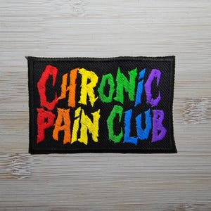 Può includere: Una toppa ricamata nera con il testo "CHRONIC PAIN CLUB" in colori arcobaleno.