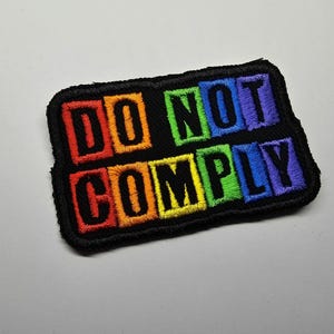Könnte beinhalten: Gestickter Aufnäher mit dem Schriftzug "DO NOT COMPLY" in Regenbogenfarben auf schwarzem Hintergrund. Der Aufnäher hat einen schwarzen Rand und eine rechteckige Form. Der Text ist in einer fetten, blockartigen Schriftart.