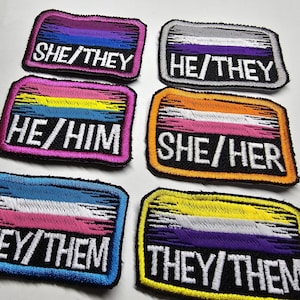 May include: Six embroidered patches with different pride flags and text. The flags are in the colors of the transgender, non-binary, bisexual, lesbian, pansexual, and genderfluid pride flags. The text on the patches reads "SHE/THEY", "HE/THEY", "HE/HIM", "SHE/HER", "THEY/THEM", and "THEY/THEM".