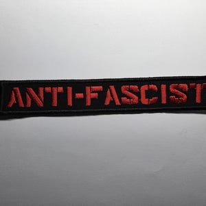May include: A rectangular black patch with the words "ANTI-FASCIST" embroidered in red. The patch is likely designed to be sewn or ironed onto clothing or fabric items.